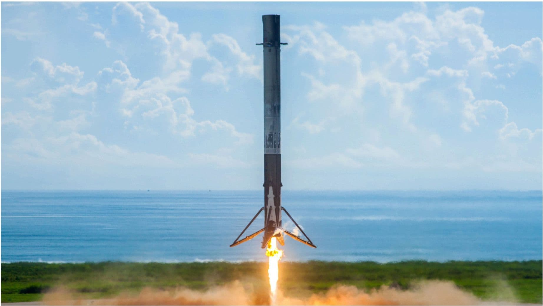 A Falcon 9 booster touching down vertically on a green coastal landing pad with the ocean horizon behind.