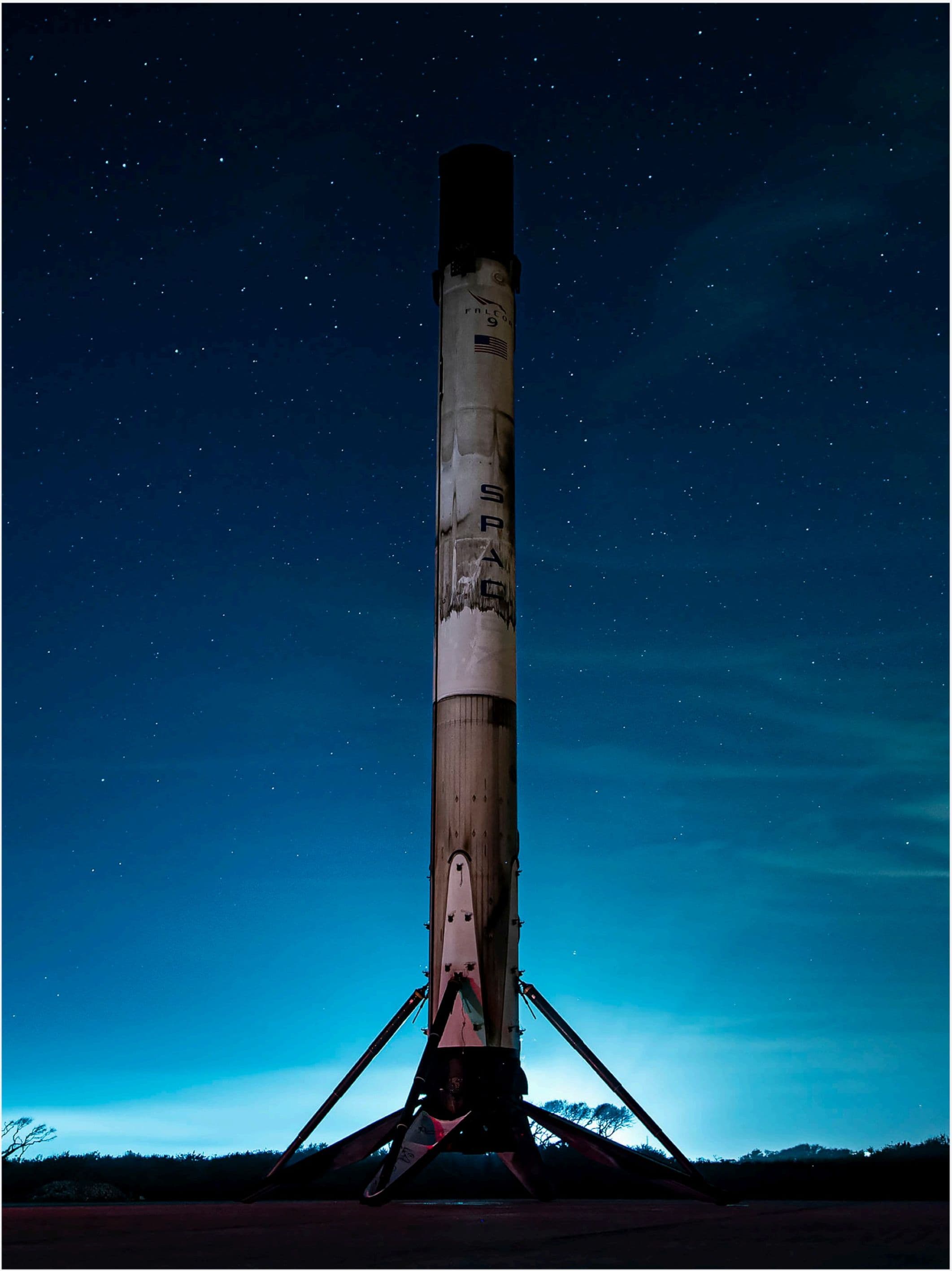 A flight-proven Falcon 9 first stage standing upright on its landing legs, scorched from reentry, beneath a star-filled twilight sky.