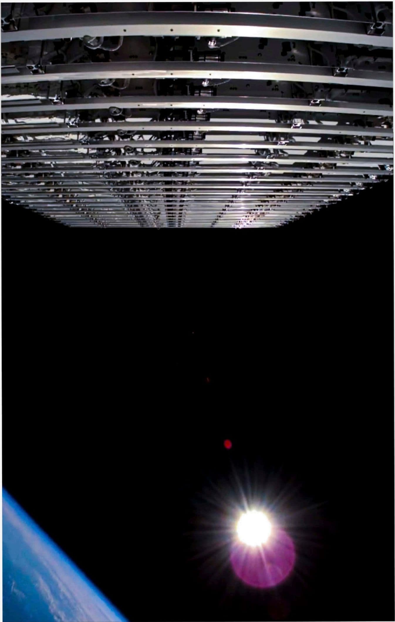View from beneath a deployed Starlink satellite stack in orbit; the edge of Earth visible at lower left, sun-flare in frame.