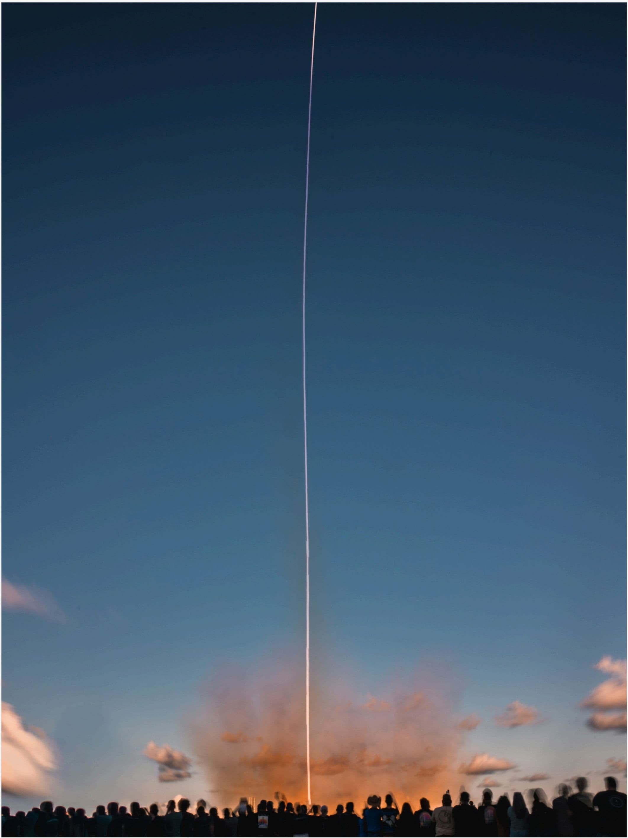 A vertical contrail rising thousands of feet against blue twilight, with silhouettes of a small crowd watching at the bottom.